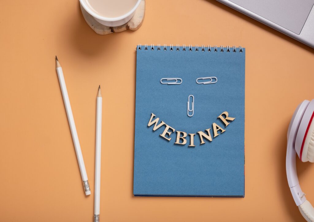 Notizblock mit Aufschrift „Webinar“ auf orangem Grund, umgeben von Laptop, Stiften und Tasse.