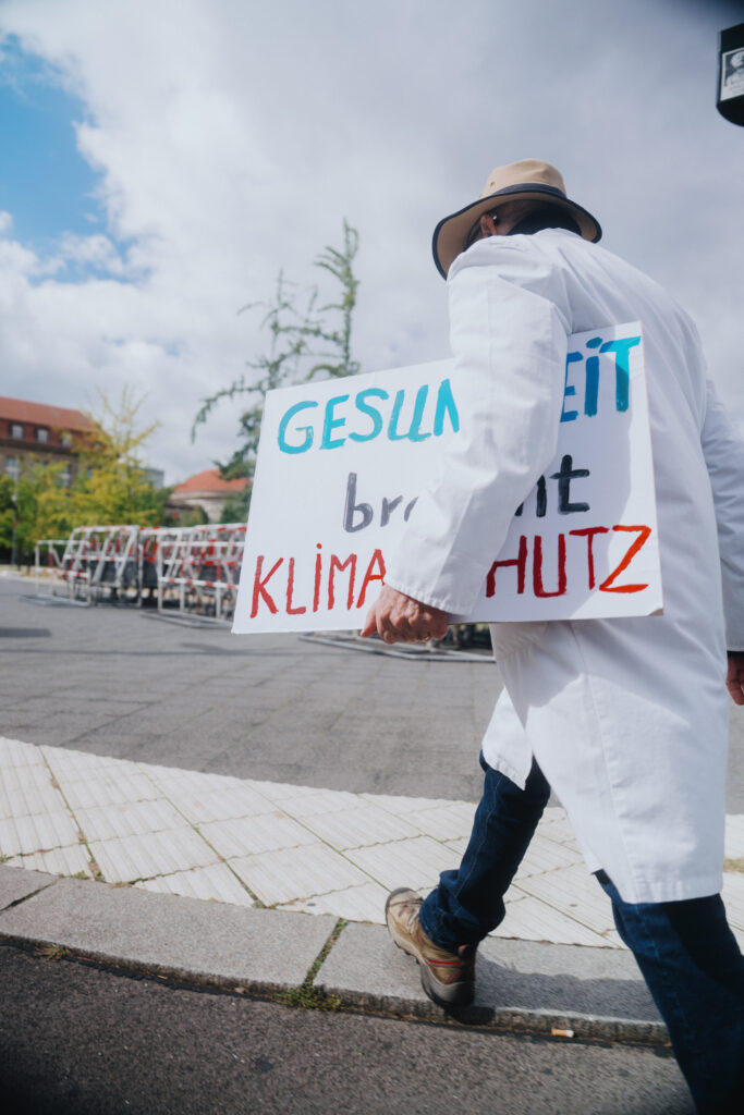 Person im weißen Kittel trägt Schild „Gesundheit braucht Klimaschutz“ auf Gehweg.