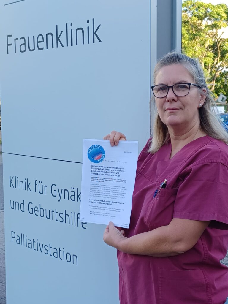 Frau in medizinischer Arbeitskleidung hält einen Brief vor einem Schild mit der Aufschrift 'Frauenklinik Klinik für Gynäkologie und Geburtshilfe Palliativstation'